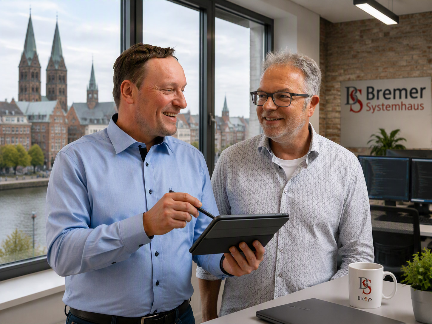 Boris Berthelmann und Ingo Langer stehen in einem Büro vor einem Fenster, hinter die Weser und den Bremer Dom sieht. Boris hält ein Tablet mit einem Stift in der Hand. Eine Tasse und ein Firmenschild mit der Aufschrift „Bremer Systemhaus“ sind zu sehen.